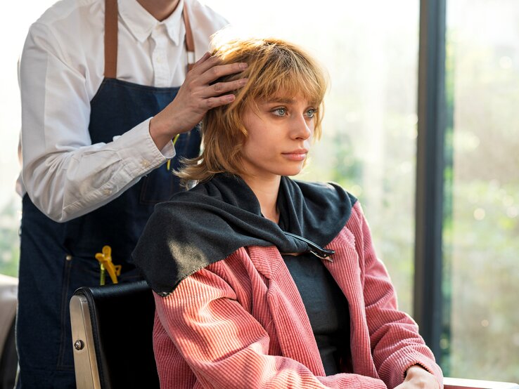 Friseur im weißen Hemd und blauer Schürze stylt Haare einer Kundin in pinkfarbenem Cordmantel vor heller Fensterfront.