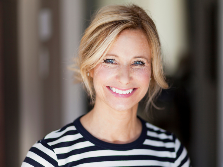 Frau mit blondem, leicht zerzaustem Haar trägt gestreiftes Shirt, aufgenommen in einem Innenraum mit verschwommenem Hintergrund.