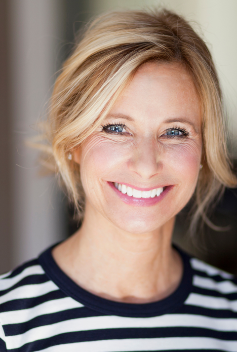 Frau mit blondem, leicht zerzaustem Haar trägt gestreiftes Shirt, aufgenommen in einem Innenraum mit verschwommenem Hintergrund.