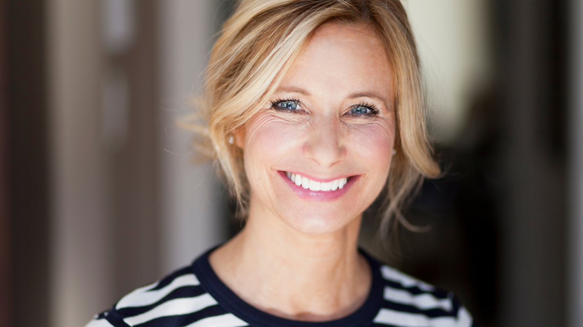 Frau mit blondem, leicht zerzaustem Haar trägt gestreiftes Shirt, aufgenommen in einem Innenraum mit verschwommenem Hintergrund.