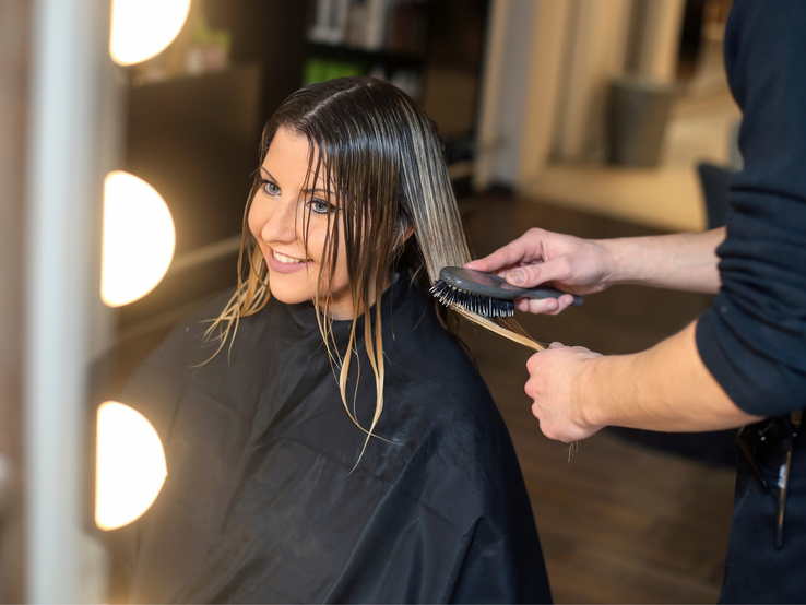 Friseur bürstet blondiertes, nasses Haar einer Person in schwarzem Umhang bei warmem Licht im modernen Salon.