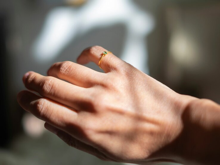Eine Nahaufnahme einer Hand, die einen kleinen Ring mit einem grünen Stein am kleinen Finger trägt. Die Hand ist im Sonnenlicht, was für einen schönen Glanz und Schattenspiel sorgt. Der Hintergrund ist unscharf, was den Fokus auf den Ring und die Hand legt."