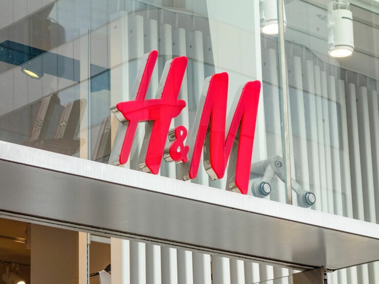 Das Bild zeigt das leuchtend rote Logo von H&M, das an einer Ladenfront befestigt ist. Das Logo ist vor einem gläsernen Hintergrund mit Sicht auf innenliegende Strukturen und Gegenstände platziert. Darüber sind moderne Beleuchtungselemente angebracht.