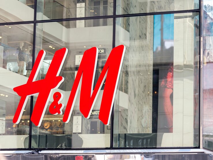 Das Bild zeigt das große, leuchtend rote Logo von H&M an der Fassade eines Geschäfts. Durch die großen Fenster hinter dem Logo sind Mannequins und Teile der Inneneinrichtung des Ladens zu erkennen.