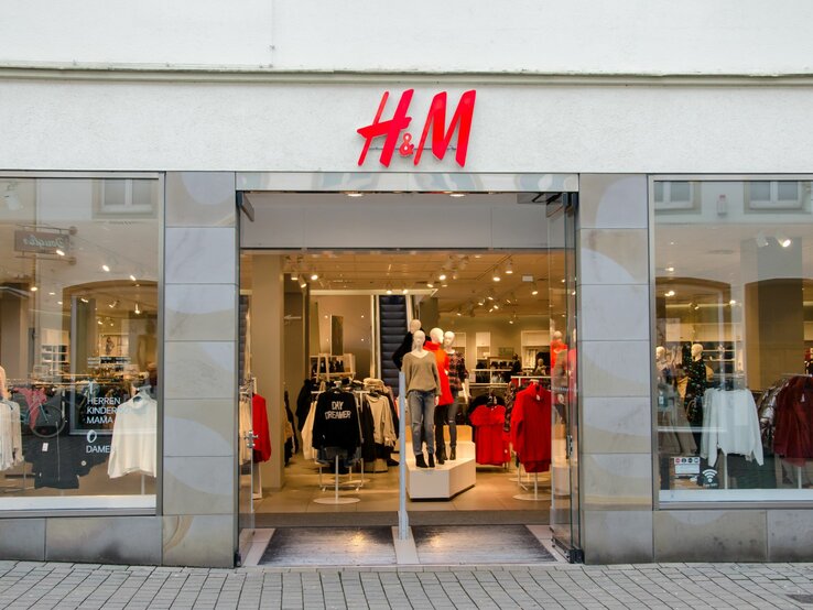 Eingang eines H&M-Geschäfts mit offenen Glastüren. Über dem Eingang befindet sich das rot-weiße H&M-Logo. Im Schaufenster sind Mannequins in aktueller Mode bekleidet, darunter ein rotes Oberteil und eine schwarze Jacke. Schilder im Inneren deuten auf verschiedene Bekleidungsabteilungen wie Herren, Kinder, Mama und Damen hin.