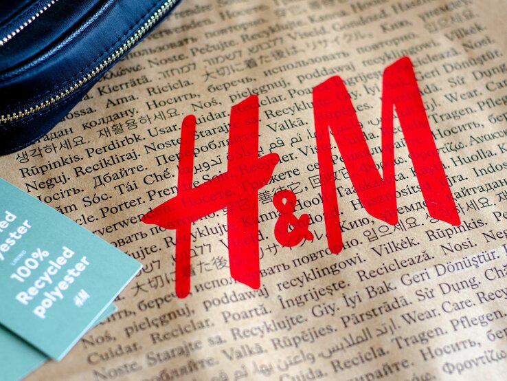 Zu sehen ist eine braune Tüte, auf der in verschiedenen Sprachen "Tragen. Pflegen. Wiederverwenden" zu lesen ist sowie in großen roten Buchstaben "H&M". Auf der linken Seite liegt ein hellgrünes Etikett mit der Aufschrift "100% Recycled Polyester" und ein Stück eines dunkelblauen Portemonnaies.