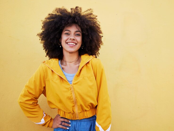 Eine fröhliche junge Frau mit voluminösem, lockigem Afrohaar steht vor einer gelben Wand. Sie trägt eine leuchtend gelbe Jacke über einem hellblauen Top, was ein lebendiges und farbenfrohes Outfit ergibt. Ihr breites Lächeln und ihre geschlossenen Augen vermitteln ein Gefühl von Entspannung und Glück. Das Bild strahlt positive Energie und sommerliche Frische aus.