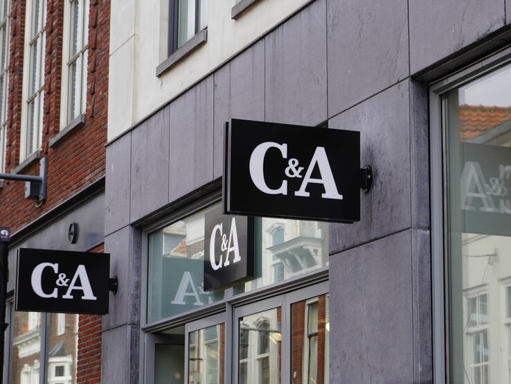 Das Foto zeigt mehrere Schilder des Bekleidungsgeschäfts C&A an der Außenfassade eines modernen Gebäudes. Die Schilder sind schwarz mit dem weißen Logo des Unternehmens darauf. Sie sind so angebracht, dass sie sowohl von Fußgängern auf der Straße als auch von weiter entfernt sichtbar sind. Im Hintergrund sieht man das typische Stadtbild mit Ziegelgebäuden und Fenstern. Das Foto vermittelt einen städtischen Eindruck und hebt die Markenpräsenz im öffentlichen Raum hervor.