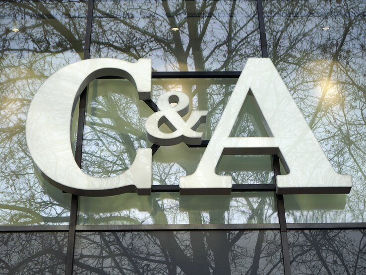 Das Foto zeigt das große weiße Logo "C&A" vor einer Fensterfront, in der sich Bäume spiegeln. Das Logo hebt sich deutlich gegen den durch die Spiegelung erzeugten natürlichen Hintergrund ab. Die Szene wirkt ruhig und die Farben sind harmonisch aufeinander abgestimmt, was einen interessanten visuellen Effekt erzeugt.