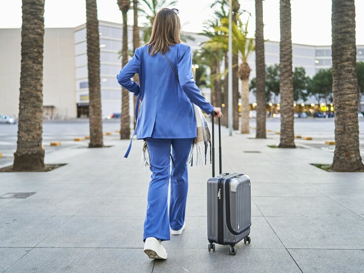 Frau in blauem Outfit von hinten mit Koffern, links und rechts Palmen
