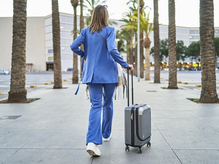 Outfits zum Reisen Frau in blauem Outfit von hinten mit Koffern, links und rechts Palmen