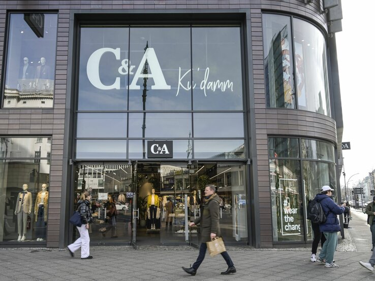 Auf diesem Foto ist eine Filiale von C&A zu sehen, welche an einem offensichtlich urbanen und belebten Standort liegt. Auffällig ist die große Beschriftung "C&A Ku'damm" auf den Fenstern, was darauf hindeutet, dass sich das Geschäft am oder in der Nähe des bekannten Kurfürstendamms in Berlin befinden könnte. Die großflächigen Schaufenster präsentieren verschiedene Modelle, die aktuelle Modekollektionen darstellen. Vorbeigehende Passanten vermitteln eine Atmosphäre der Geschäftigkeit, was typisch für eine Einkaufsstraße in einer Großstadt ist. Das Foto fängt das dynamische Treiben des städtischen Alltags ein und zeigt gleichzeitig die Markenpräsenz von C&A im Einzelhandel.