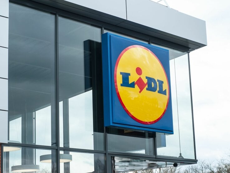  Das Bild zeigt das typische Logo des Einzelhändlers Lidl an einem Geschäftsgebäude. Die moderne Architektur des Gebäudes mit großen Glasflächen und der klare Himmel im Hintergrund vermitteln einen zeitgenössischen Eindruck. Lidl ist bekannt für sein Discount-Supermarktkonzept, das in vielen Ländern vertreten ist und eine breite Palette von Produkten, darunter Lebensmittel, Haushaltswaren und saisonale Artikel, zu günstigen Preisen anbietet. Das präsentierte Logo mit dem blauen Hintergrund und dem gelben Kreis mit roter Schrift ist sofort erkennbar und repräsentiert die Marke Lidl weltweit.