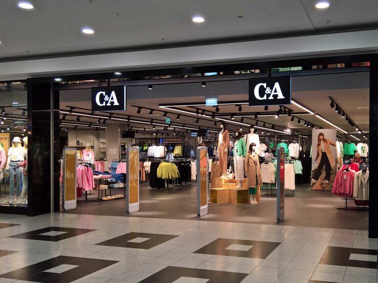 Der Eingangsbereich einer C&A-Filiale in einem Einkaufszentrum. Über dem Eingang hängt ein großes, beleuchtetes C&A-Logo. Im Inneren des Geschäfts sind mehrere Kleidungsständer und Schaufensterpuppen zu sehen, die aktuelle Mode präsentieren. Die Schaufensterpuppen tragen verschiedene Outfits, von Freizeitkleidung bis zu eleganteren Stücken. An der rechten Seite des Eingangs ist ein großes Werbeplakat mit einem Model in einem trendigen Outfit. Der Boden des Einkaufszentrums ist mit dunklen und hellen Fliesen gemustert, und die Beleuchtung im Laden wirkt einladend.