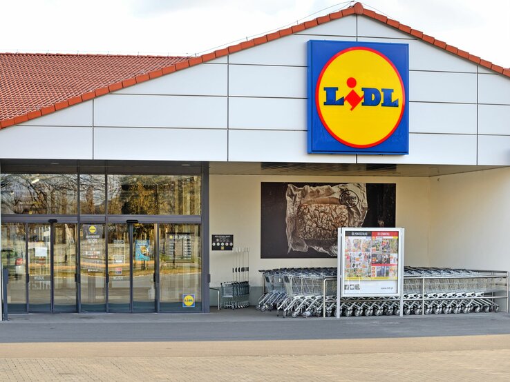 Auf dem Foto ist der Eingangsbereich einer Lidl-Filiale zu sehen. Über dem Eingang prangt das charakteristische Firmenschild mit gelbem Kreis und dem Lidl-Logo in der Mitte. Das Gebäude hat ein schlichtes Design mit einer weißen Fassade und einem roten Ziegeldach. Vor dem Eingang sind Glasautomatiktüren und eine Reihe von Einkaufswagen. An der linken Seite des Eingangs befindet sich ein Plakat mit Werbung für Produkte, die im Laden erhältlich sind. Im Hintergrund sind Bäume und ein parkähnlicher Bereich zu erkennen.