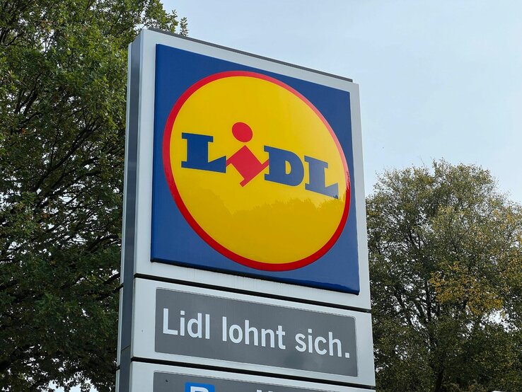 Auf dem Bild ist ein Werbeschild von Lidl zu sehen. Im oberen Teil des Schildes ist das typische Lidl-Logo mit gelbem Hintergrund und dem roten Lidl-Schriftzug. Unterhalb des Logos befindet sich der Slogan „Lidl lohnt sich.“ im Weiß auf einem blauen Hintergrund. Im Hintergrund des Schildes sind Bäume mit grünem Laub zu erkennen, was auf eine Aufnahme bei Tageslicht hindeutet.