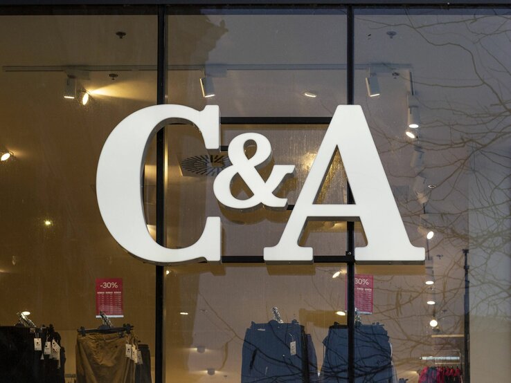  Das Foto zeigt das große, weiße Logo von C&A, das an der Innenseite einer Fensterfront eines Geschäfts angebracht ist. Man kann durch das Fenster in den Laden blicken und einige Kleidungsstücke und Verkaufsschilder mit Rabattangaben sehen, die im Inneren ausgestellt sind. Die Reflexion der Bäume auf der Fensterscheibe lässt darauf schließen, dass das Foto von außen bei Tageslicht aufgenommen wurde.