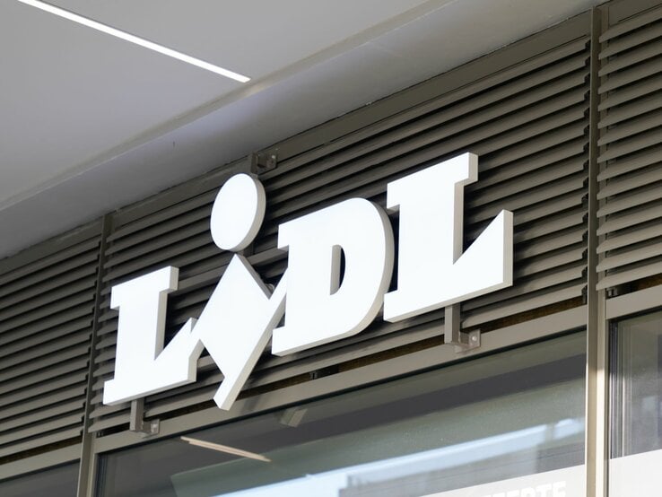 Das Foto zeigt das weiße 3D-Schriftzug-Logo von Lidl, angebracht an einer Ladenfassade über einem Eingang. Das Logo ist vor einem Hintergrund aus horizontalen Lamellen platziert, die typisch für Ladenfronten sind, um Sonnenlicht zu filtern und für Privatsphäre zu sorgen. Die Aufnahme wirkt modern und ist vermutlich tagsüber gemacht worden, wobei das Logo durch seine erhöhte Montage und Farbgebung einen markanten visuellen Akzent setzt.