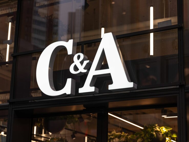  Auf dem Foto ist das beleuchtete Logo des Modeunternehmens C&A zu sehen, das an einem Fenster eines Geschäfts befestigt ist. Die großen, weißen Buchstaben heben sich deutlich gegen die dunkle Umgebung ab. Hinter dem Logo ist eine Innenansicht des Geschäfts mit Pflanzen und einer Reihe von Lichtern zu erkennen, die für eine warme und einladende Atmosphäre sorgen.