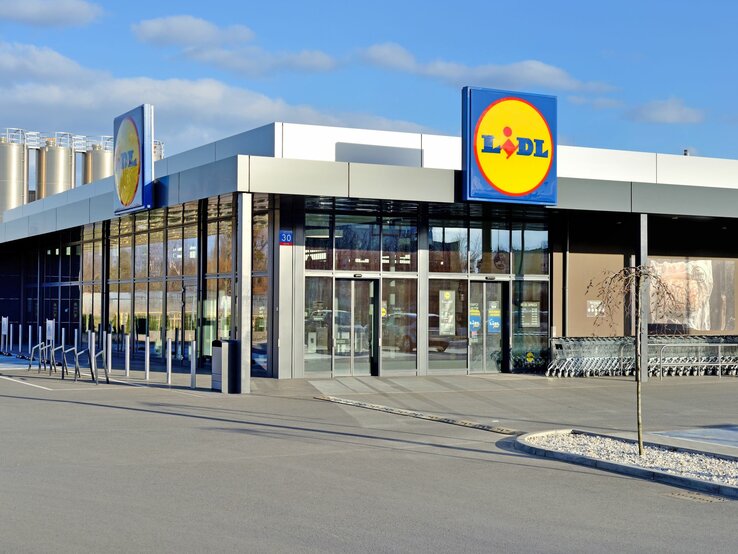 Auf dem Bild ist eine moderne Lidl-Filiale zu sehen. Sie hat eine schlichte und zeitgenössische Architektur mit einer klaren Glasfront und einem flachen Dach. Über dem Eingang ist ein großes, beleuchtetes Lidl-Logo angebracht. Vor dem Supermarkt befinden sich ordentlich aufgereihte Einkaufswagen. Im Hintergrund sind silberfarbene Silos zu sehen, was auf eine industrielle Umgebung hindeutet. Die Szene ist bei Tageslicht aufgenommen, mit einem klaren blauen Himmel im Hintergrund.