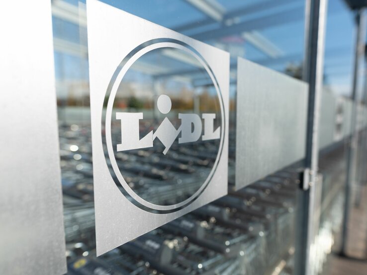 Das Foto zeigt das Logo von Lidl auf einer Glasscheibe, wahrscheinlich am Eingang eines Lidl-Marktes. Das Logo ist mattiert auf der durchsichtigen Fläche, und im Hintergrund sind verschwommen Einkaufswagen zu erkennen, typisch für den Außenbereich eines Supermarktes. Die Aufnahme spielt mit der Tiefenschärfe und fokussiert das Logo, während die Umgebung unscharf bleibt.