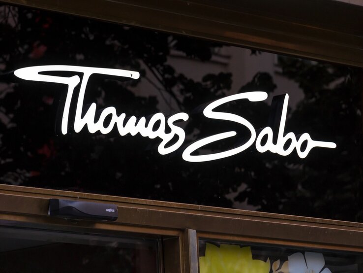 Eleganter Thomas Sabo Schriftzug leuchtet in Weiß auf einem Ladenfenster, Bäume spiegeln sich.