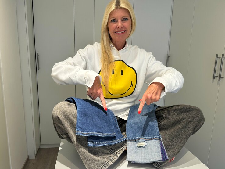 Renate Zott trägt lächelnd einen weißen Pullover mit einem großen, gelben Smiley-Gesicht darauf und zeigt auf zwei verschiedene Jeansstoff-Muster, die sie auf ihren Knien platziert hat.