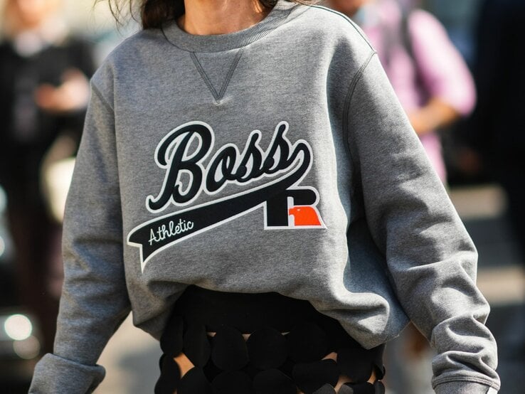 Nahaufnahme einer Frau, die ein graues Sweatshirt trägt. Auf der Vorderseite des Sweatshirts ist in Großbuchstaben das Wort "Boss" zusammen mit dem Zusatz "Athletic" aufgedruckt. Ein markantes Detail ist das eingenähte V-förmige Dreieck am Kragen, das typisch für viele Sweatshirt-Designs ist.