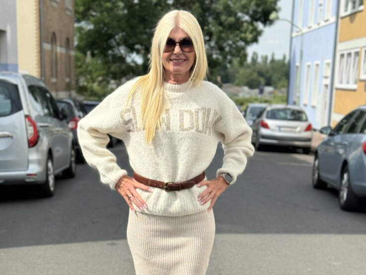 Frau mittleren Alters in einem urbanen Straßenszenario, trägt einen cremefarbenen Strickpullover mit der Aufschrift "LEAD DUKE", dazu passenden langen Strickrock und braune Stiefel. 