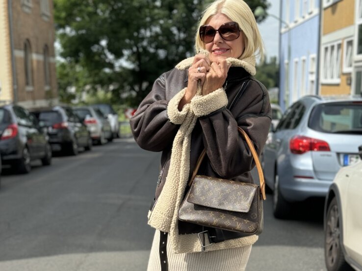 Frau mittleren Alters auf einer städtischen Straße, trägt einen dunkelbraunen Mantel mit beigefarbenem Teddyfell am Kragen und eine passende Handtasche. Sie hat einen langen, beigen Strickrock an, kombiniert mit braunen Stiefeln.