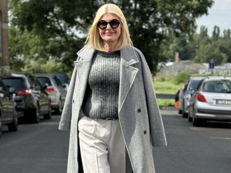 Porträt einer eleganten, älteren Frau, die auf einer städtischen Straße läuft. Sie trägt eine weiße, weit geschnittene Hose, einen grauen Wollpullover und darüber einen offen getragenen grauen Mantel.