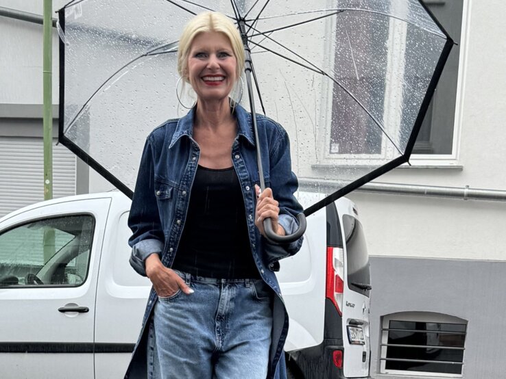 Frau mit blonden Haaren, die einen durchsichtigen Regenschirm hält. Sie trägt einen langen, offenen Jeansmantel über einem schwarzen Oberteil und einer weiten hellen Jeans.