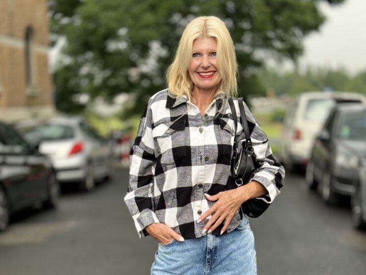Frau mit blonden Haaren, die in einem lässigen, karierten Hemd in Schwarz-Weiß und einer hellen Jeans gekleidet ist. 