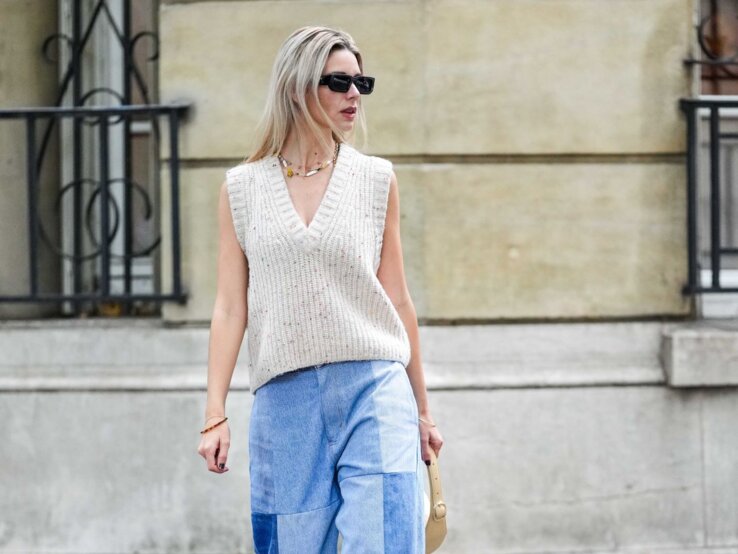 Jeans-Trend im Winter Junge blonde Frau in beigefarbenem Stricktop und weiter Patchwork-Jeans geht mit kleiner Tasche auf herbstlicher Straße.
