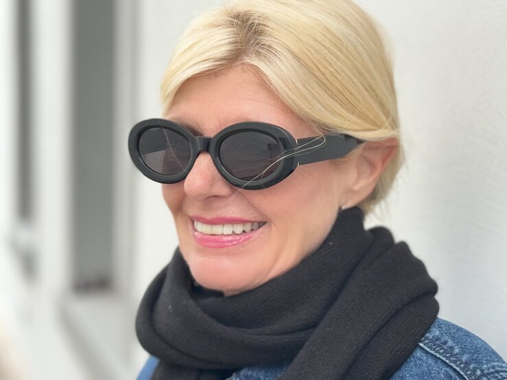 Nahaufnahme einer Frau, die eine auffällige, ovale Sonnenbrille trägt. Die Sonnenbrille hat breite, schwarze Rahmen und verleiht ihrem Look eine moderne, stilbewusste Note.