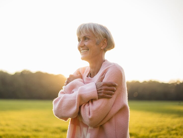 Himbeer Pullover Tchibo Ältere Frau mit grauem Haar und rosa Pullover umarmt sich lächelnd im Sonnenlicht auf einer Wiese.