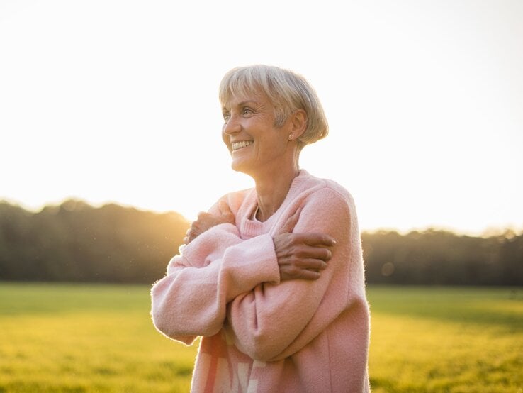 Himbeer Pullover Tchibo Ältere Frau mit grauem Haar und rosa Pullover umarmt sich lächelnd im Sonnenlicht auf einer Wiese.