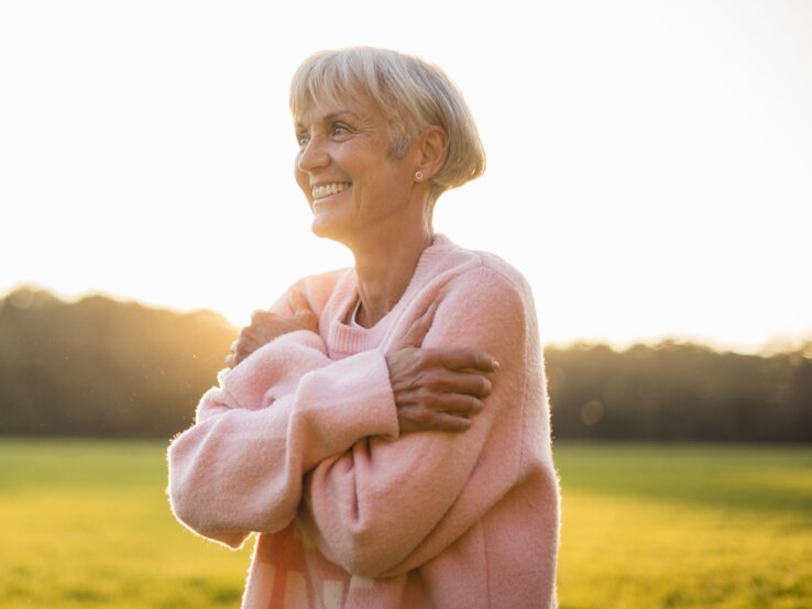 Himbeer Pullover Tchibo Ältere Frau mit grauem Haar und rosa Pullover umarmt sich lächelnd im Sonnenlicht auf einer Wiese.