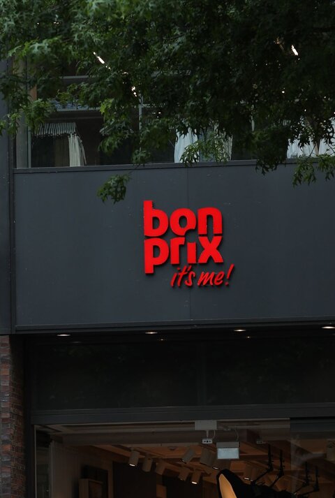 Bonprix-Logo in leuchtendem Rot auf einer grauen Fassade, daneben eine historische Steinskulptur vor Backsteinmauer.