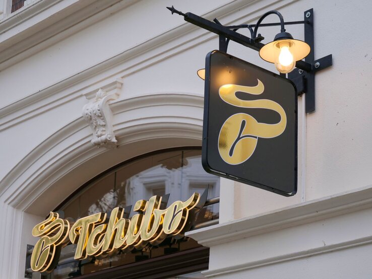 Tchibo-Schild Licht Außenansicht eines Tchibo-Geschäfts mit goldener Leuchtschrift und stilisiertem Kaffeemotiv auf schwarzem Schild.