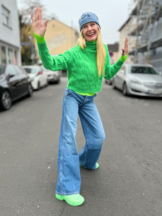 Frau mit langem blondem Haar, leuchtend grünem Pullover und grünen Schuhen macht eine spielerische Pose auf einer ruhigen Straße | © Renate Zott