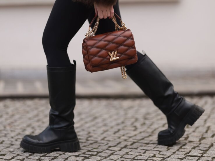 Nahaufnahme von Beinen in schwarzen Stiefeln auf Kopfsteinpflaster, eine braune Handtasche mit goldener Kette wird getragen.