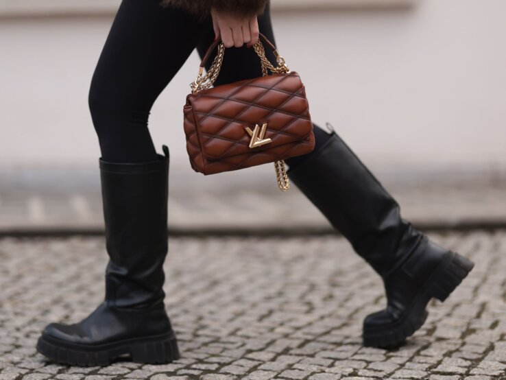 Nahaufnahme von Beinen in schwarzen Stiefeln auf Kopfsteinpflaster, eine braune Handtasche mit goldener Kette wird getragen.