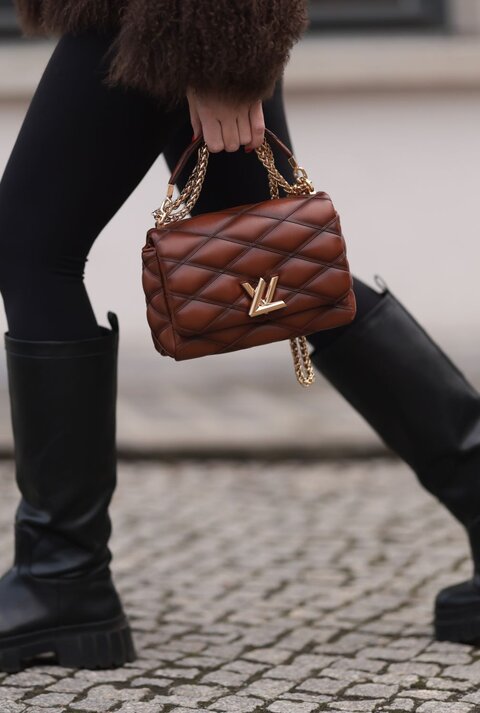 Nahaufnahme von Beinen in schwarzen Stiefeln auf Kopfsteinpflaster, eine braune Handtasche mit goldener Kette wird getragen.
