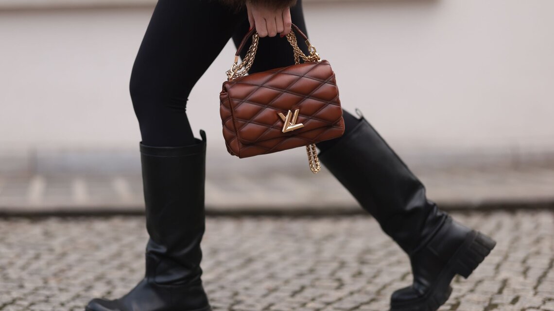 Nahaufnahme von Beinen in schwarzen Stiefeln auf Kopfsteinpflaster, eine braune Handtasche mit goldener Kette wird getragen.