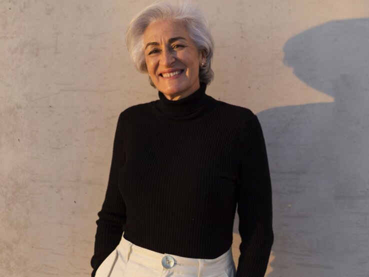 Elegante Frau mit grauem Haar in schwarzem Rollkragenpullover und heller Hose lehnt lächelnd vor einer Betonwand mit weichem Schatten.