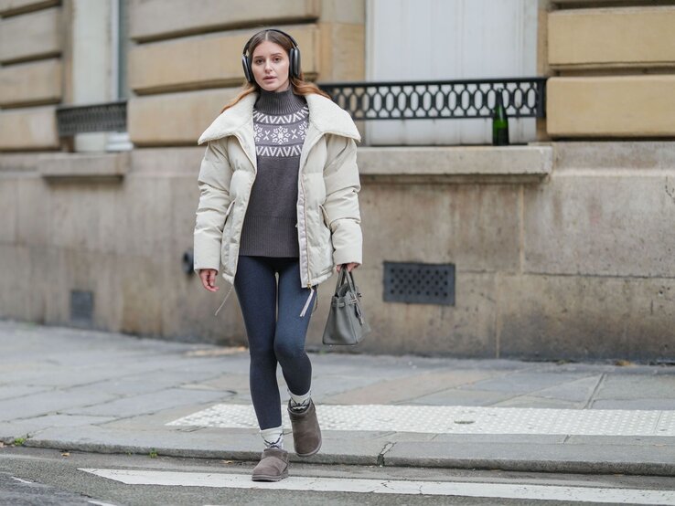 Thermoleggings Tchibo Winter Frau mit beigem Wintermantel, grauem Pullover mit Schneeflockenmuster und Thermoleggings läuft konzentriert über einen Gehweg.