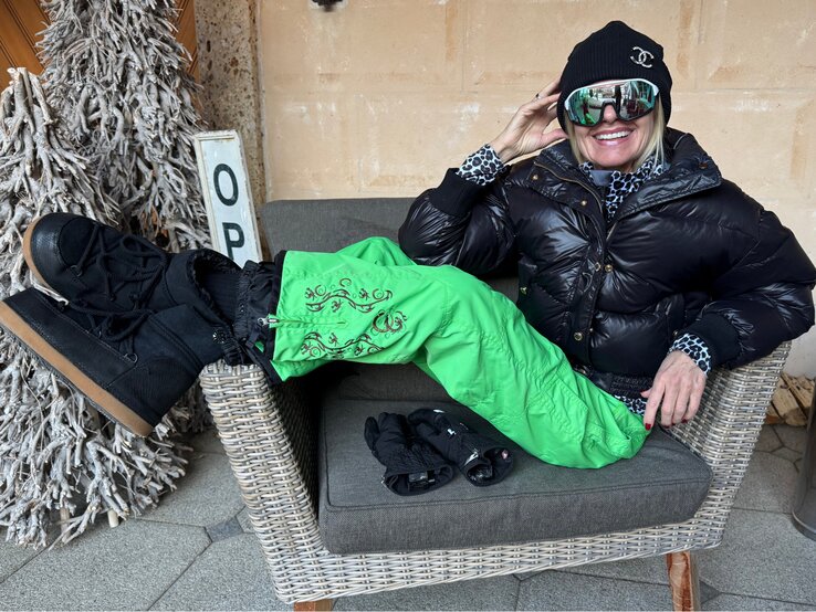 Renate Zott Ski-Outfits Renate Zott in schwarzer Daunenjacke, grüner Schneehose und dicker Mütze sitzt leger auf einem Korbstuhl, ein Handschuhpaar liegt daneben.