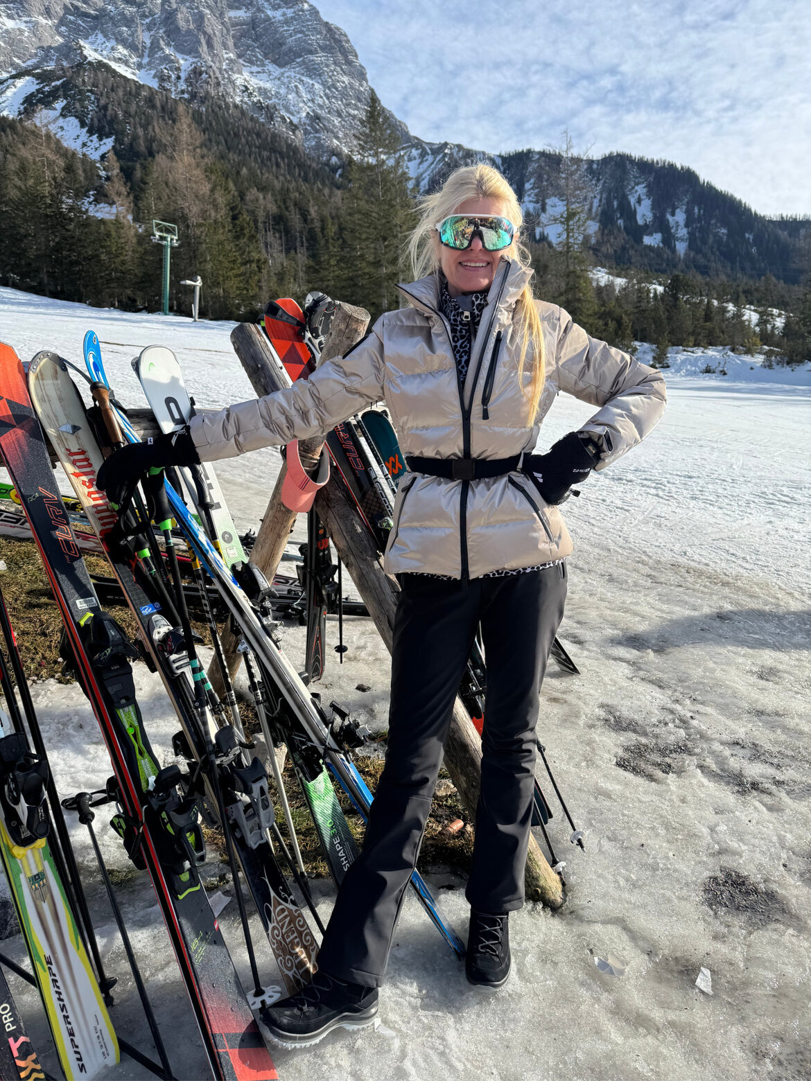 Renate Zott Ski-Outfits Metallic | © Renate Zott Renate Zott in silberner Daunenjacke, schwarzer Skihose und verspiegelter Skibrille lehnt lächelnd an einem Holzständer mit Skiern. | © Renate Zott