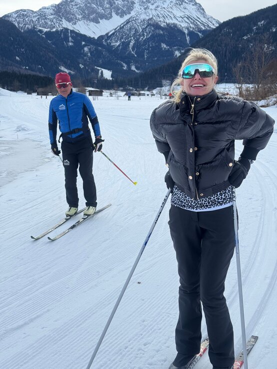 Renate Zott Ski-Langlauf mit Lehrer | © Renate Zott Renate Zott in schwarzer Daunenjacke und verspiegelter Skibrille steht lächelnd mit Langlaufskiern auf einer Schneeloipe vor Bergkulisse. | © Renate Zott